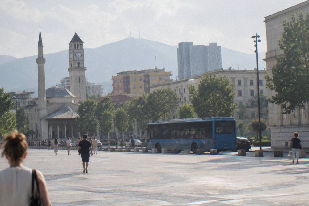 A blue Tirana city bus on a street with colourful buildings in the background