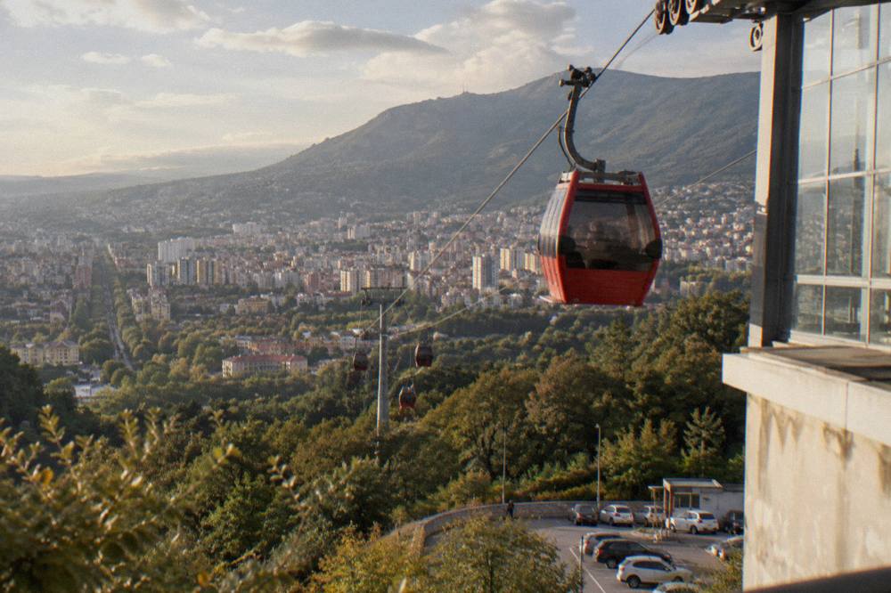 A red cable car cabin of the Dajti Ekspres rising above trees toward the mountain with the city basin visible behind