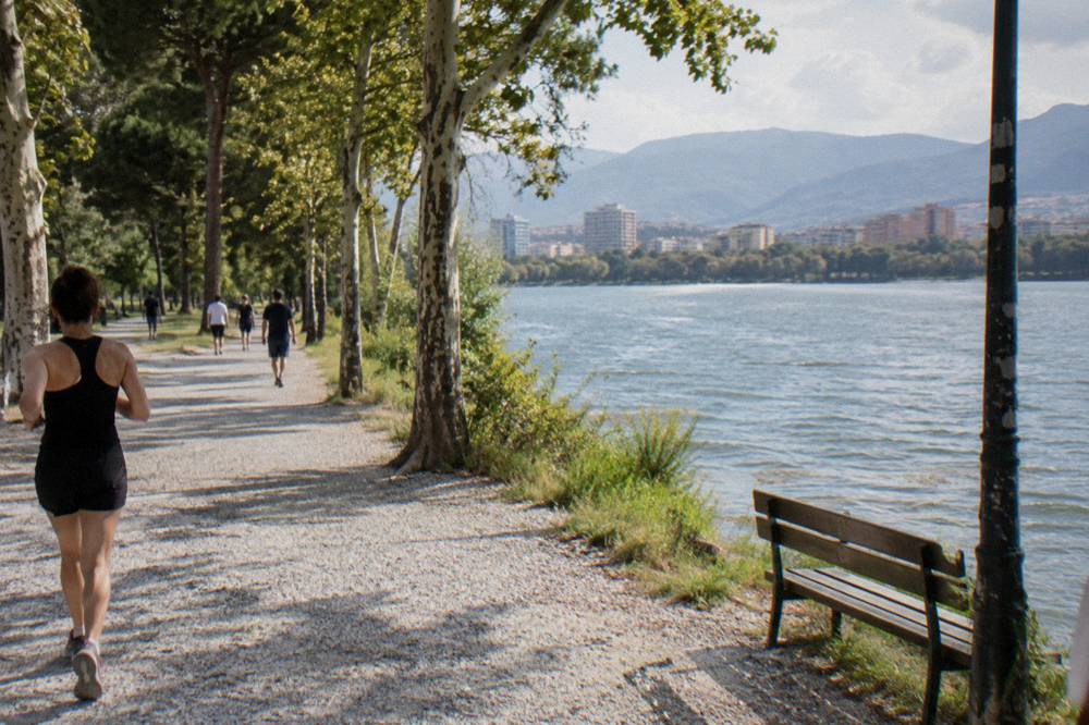 A wide public park path beside a lake with trees and people strolling on a warm afternoon