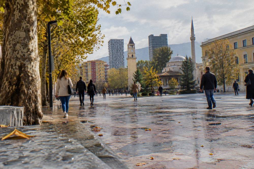 Visitors in light jackets walking a tree-lined boulevard in Tirana with autumn leaves on the pavement