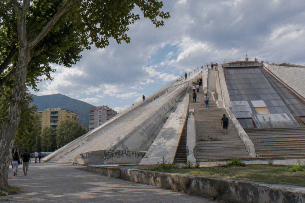 The grey concrete facade of the Pyramid of Tirana with stepped sides and people on the stairs