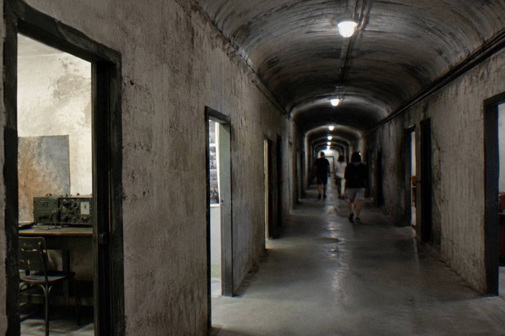 A long dim concrete corridor inside a bunker museum with rooms opening off both sides